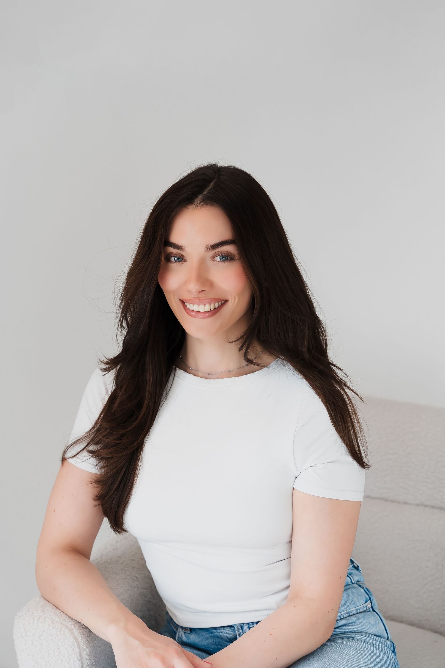 Smiling brunette woman in white t-shirt and jeans sitting relaxed on a light gray chair against white background.