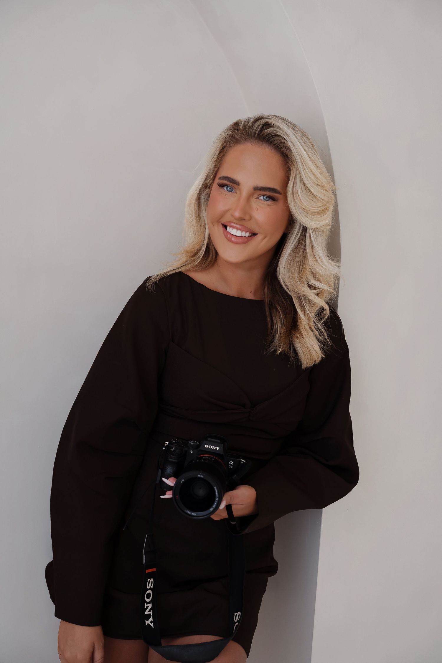 Person in black sweater holds camera while posing against white wall.