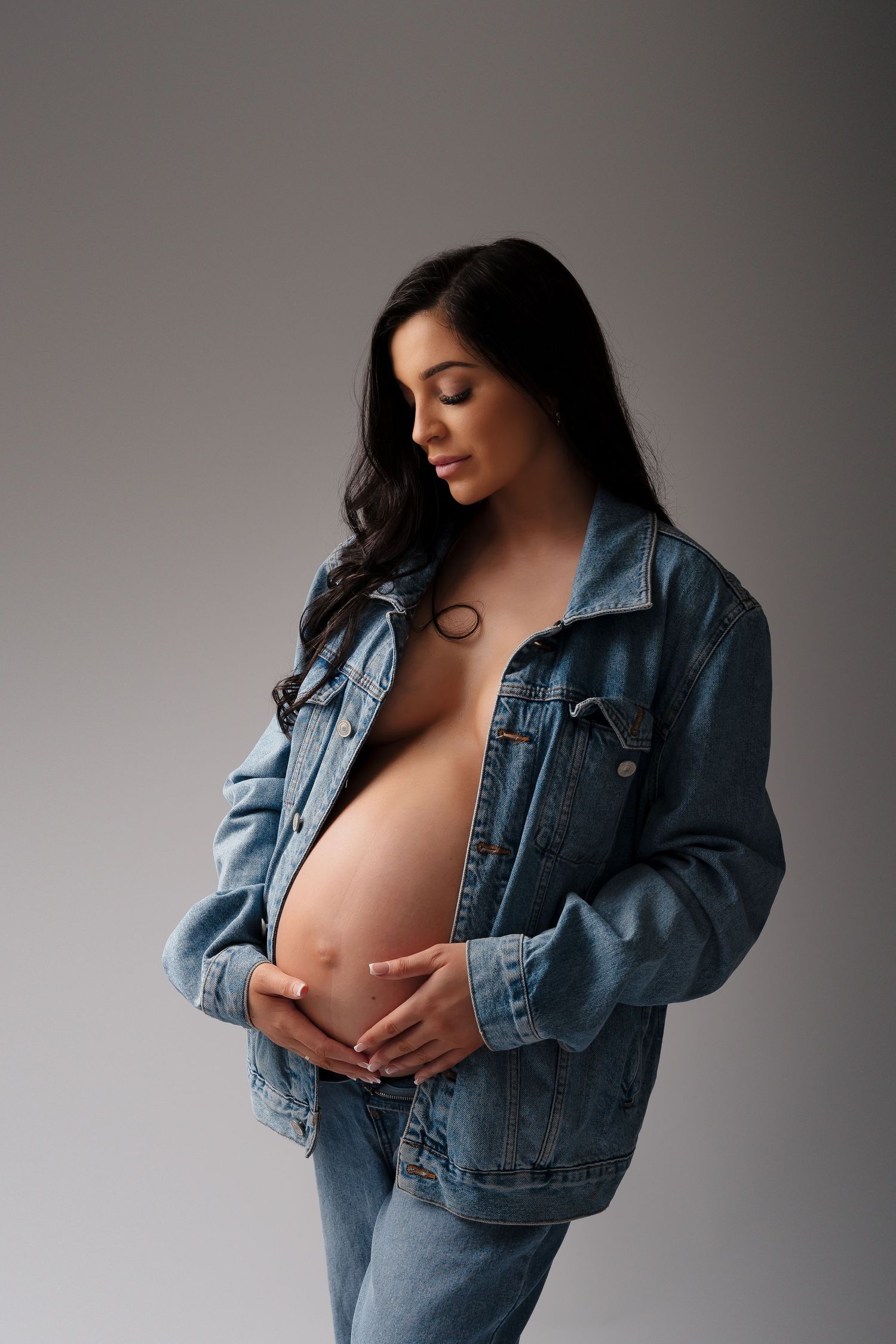 Casual maternity portrait in denim jacket showing bare belly against grey backdrop.