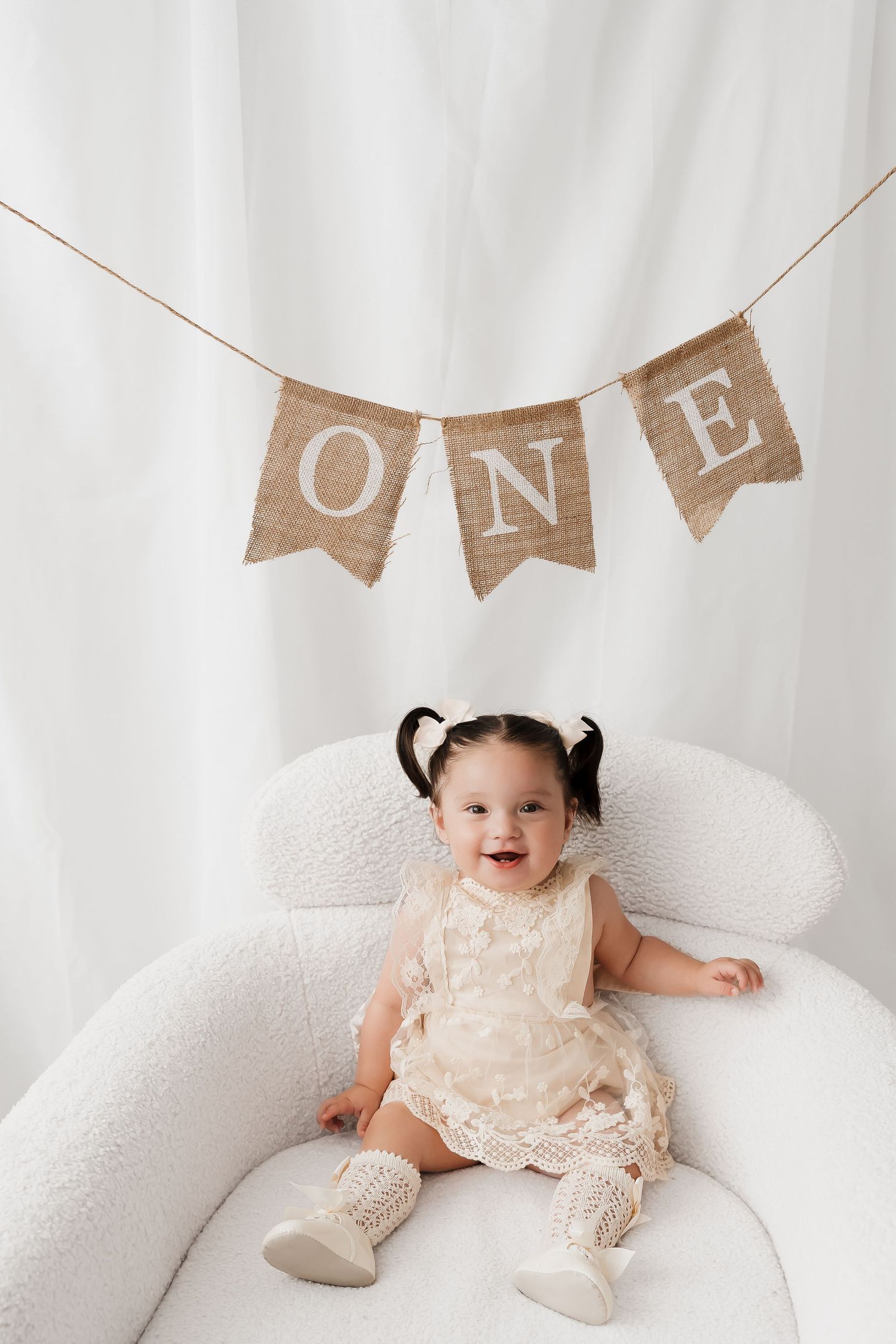Smiling baby girl in lace dress sitting under burlap ONE banner decoration.