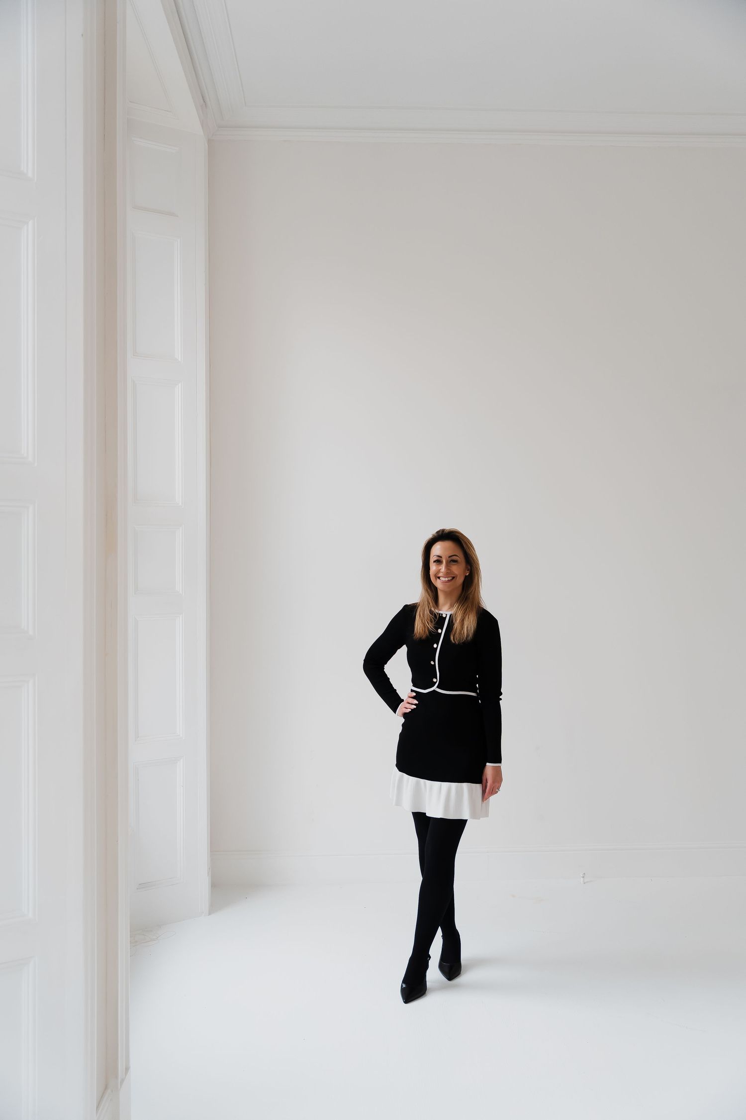Woman in black dress with white trim and black tights poses confidently in a bright white minimalist room.