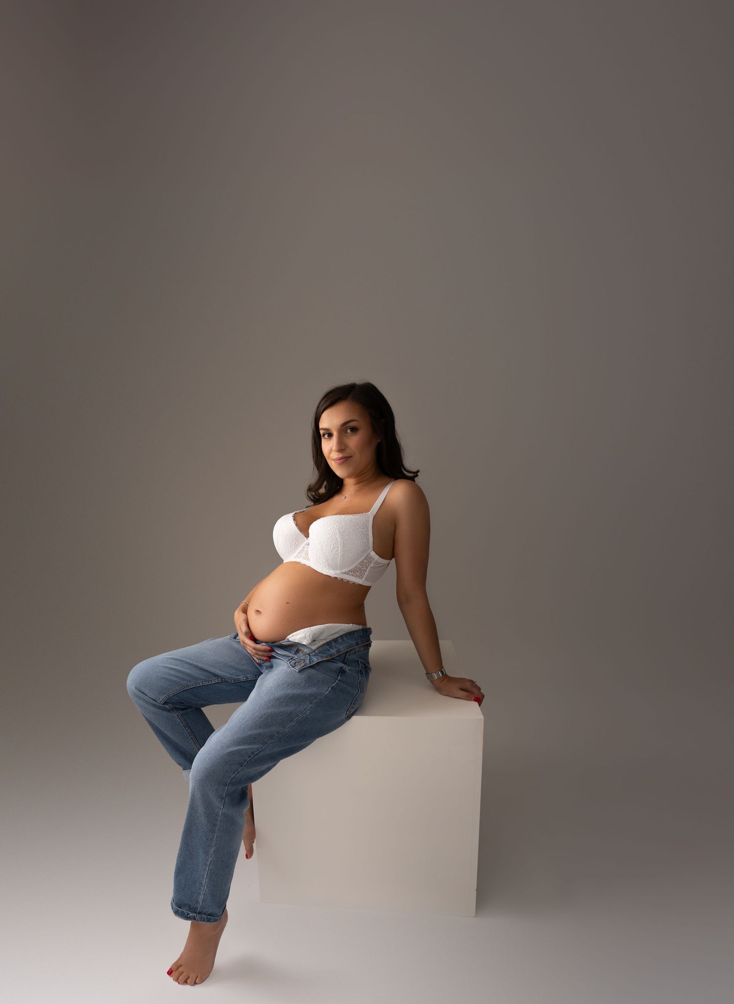 Casual maternity portrait with woman in jeans and white top sitting on white cube.
