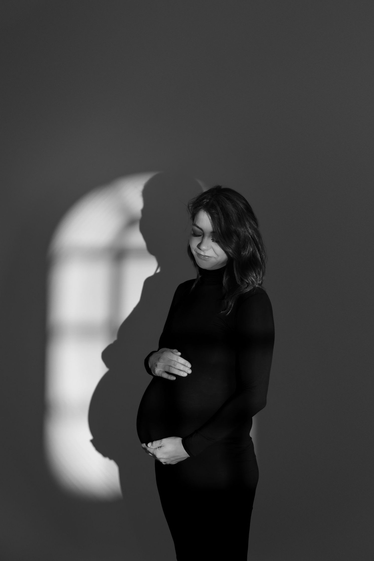 Dramatic monochrome portrait of an expectant mother with window light casting artistic shadows.