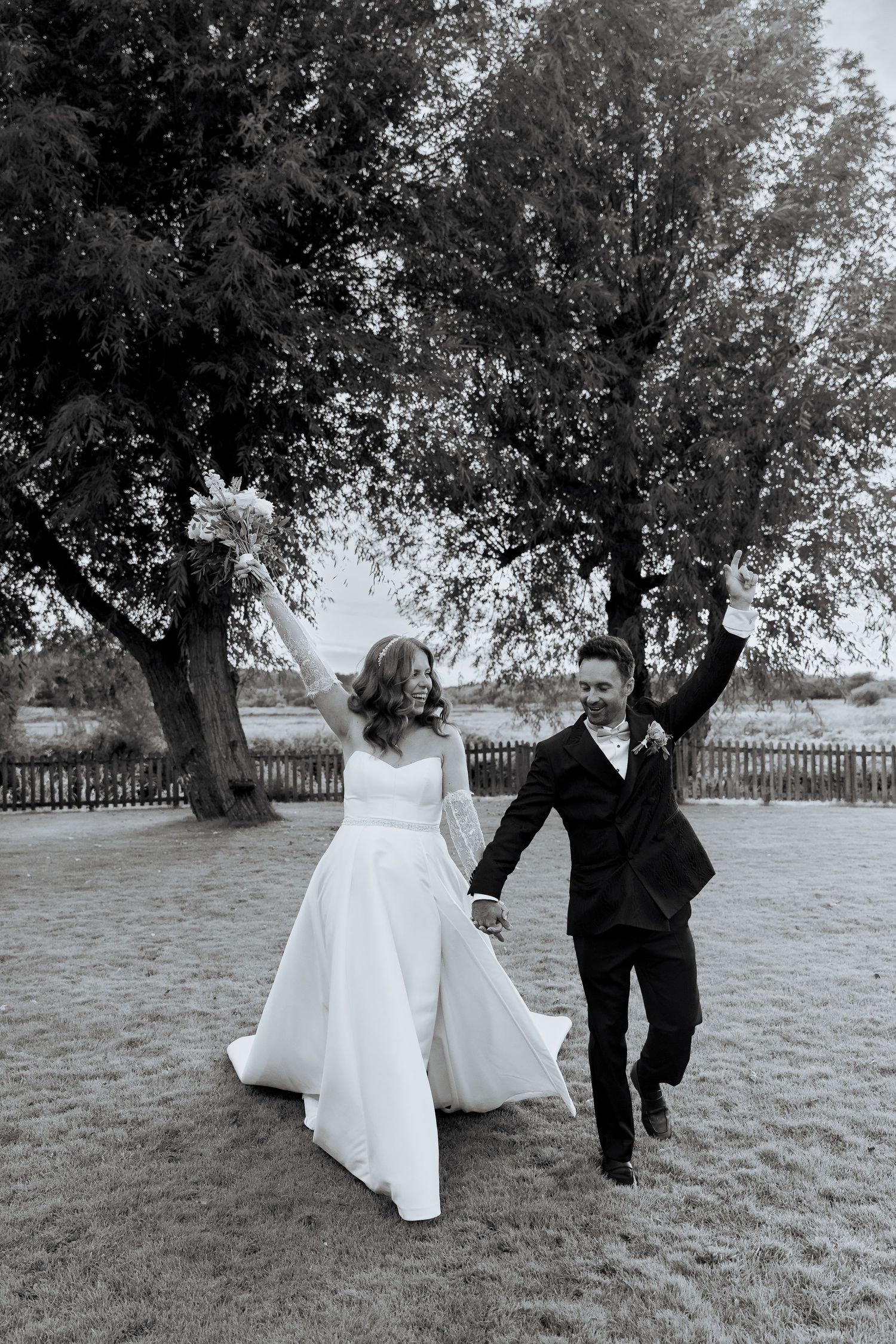 A bride and groom walking together through a field with trees in black and white.