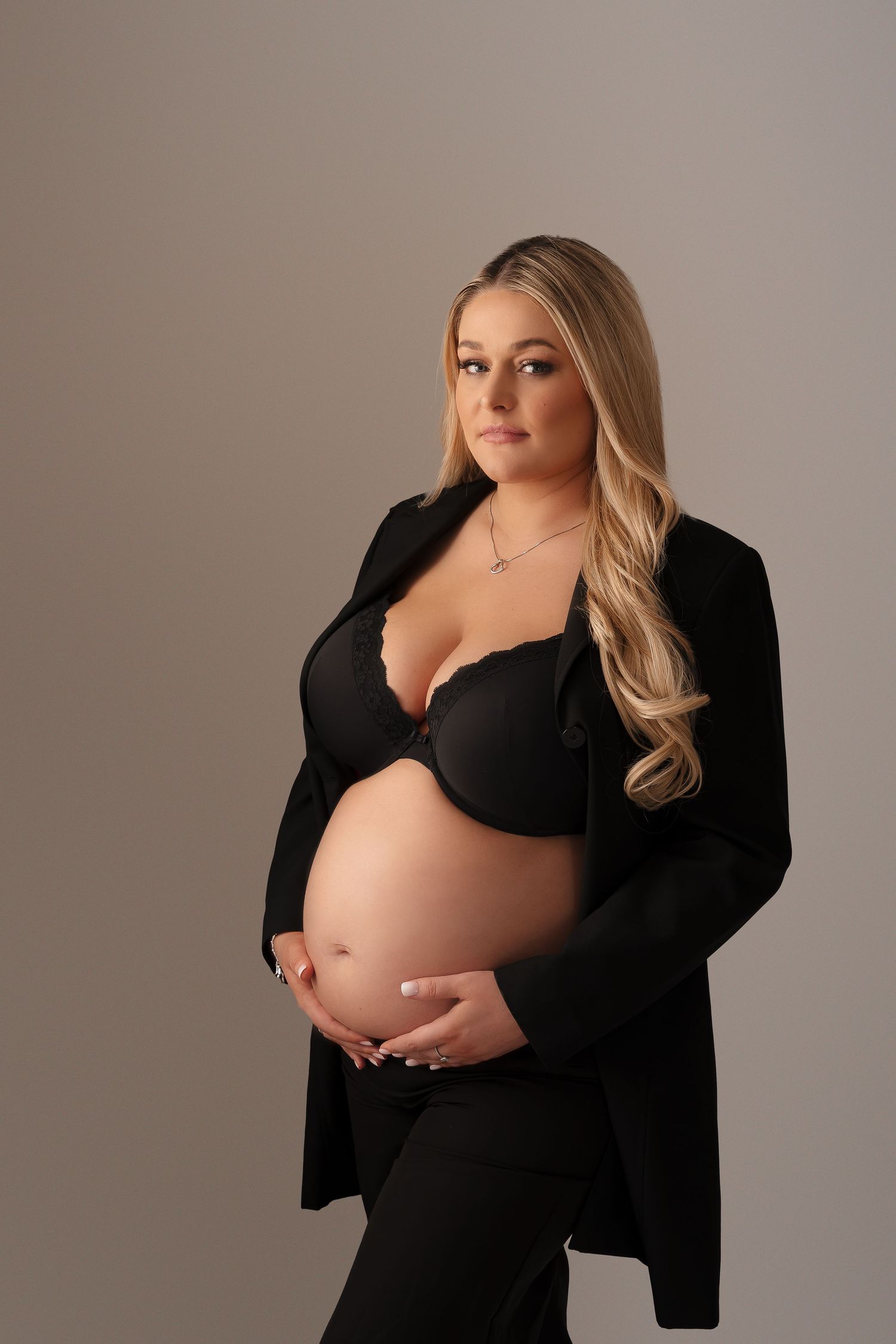 Elegant maternity portrait featuring a blonde woman in all black against a neutral backdrop.