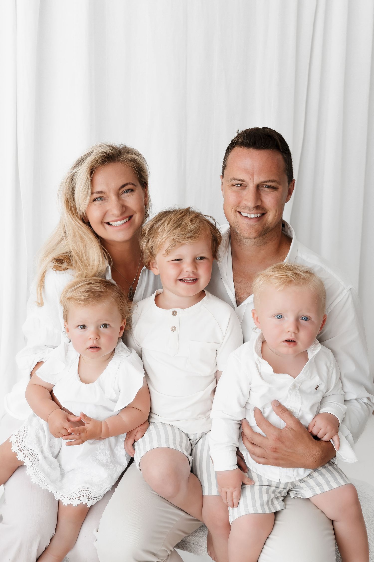 Happy family in white clothing poses together for portrait against white backdrop.