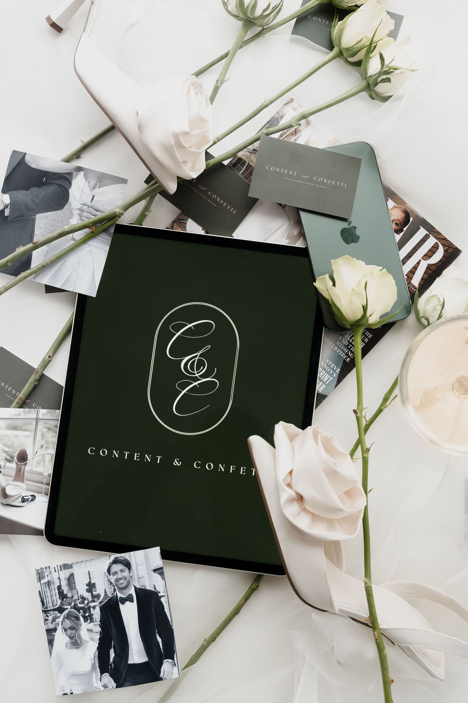 Elegant flat lay with a tablet displaying Content & Confetti logo, white roses, business cards, and wedding photos.