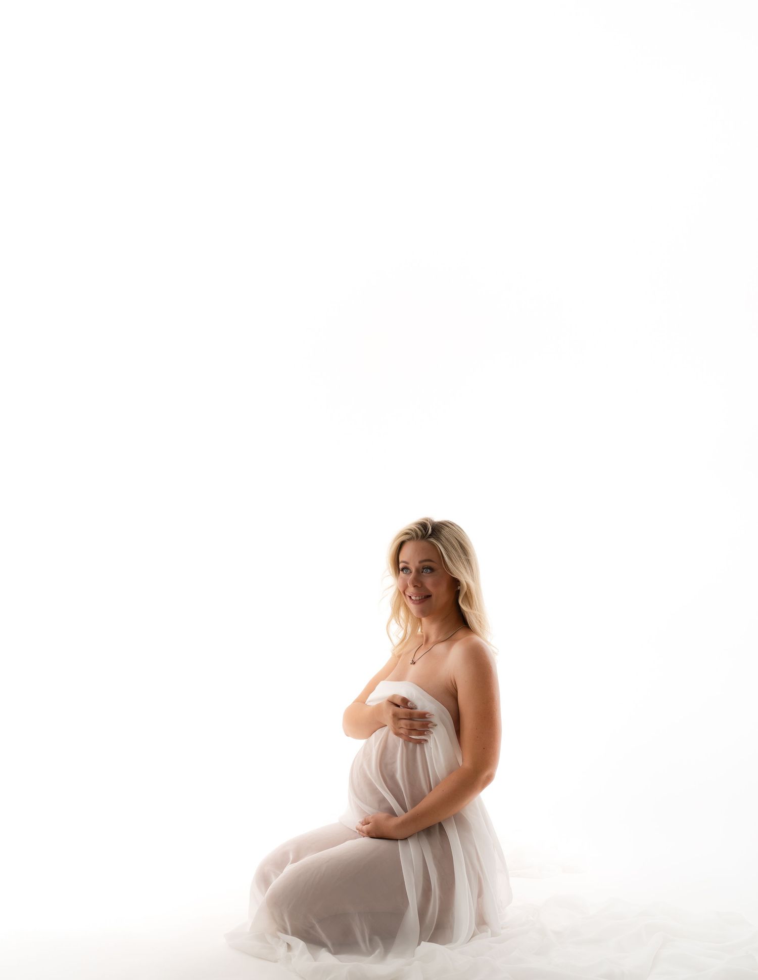 Ethereal maternity portrait with woman in flowing white dress against white background.