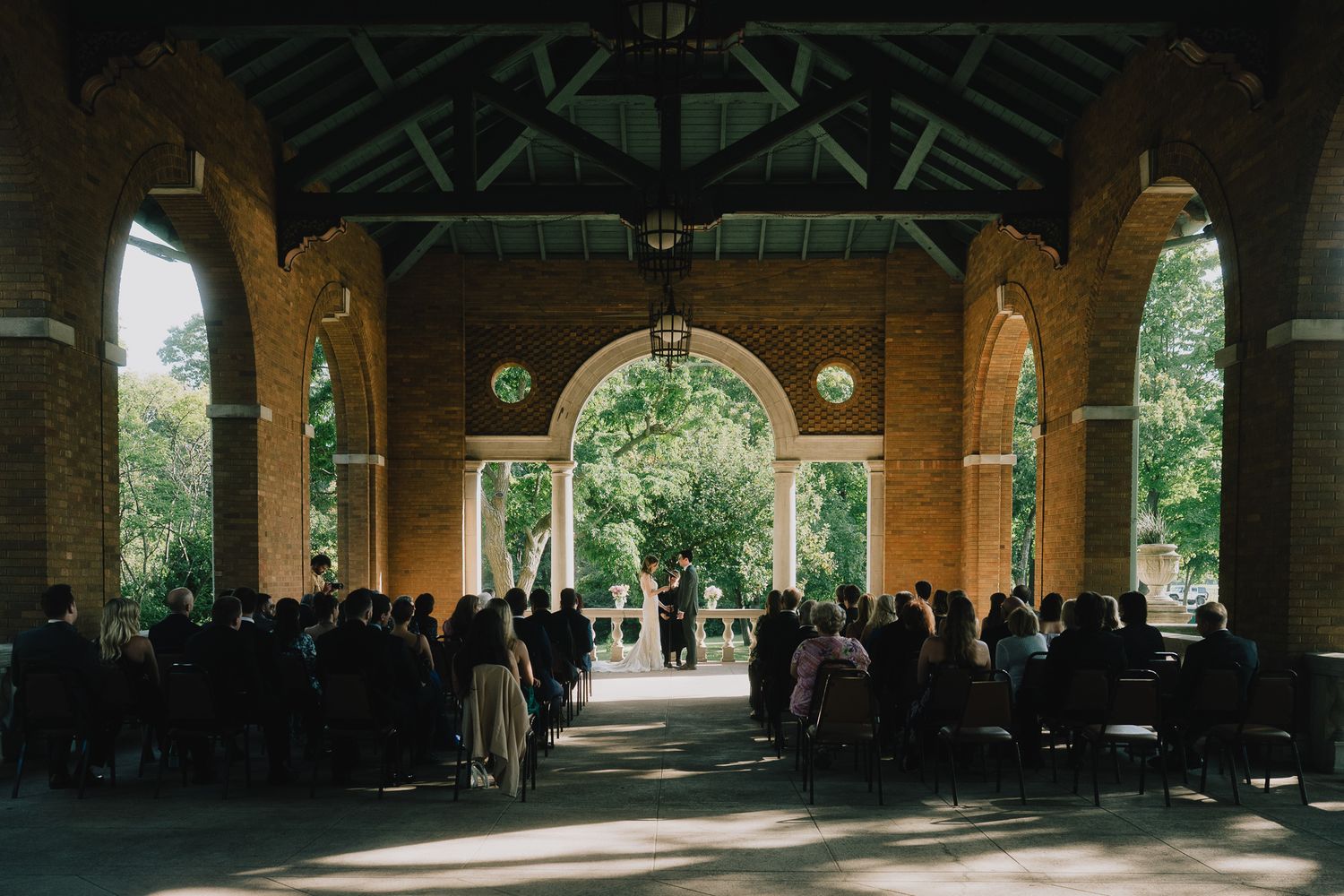 A covered outdoor wedding venue with wooden beams and stone columns in a park-like setting, with the bride and groom and wedding guests.