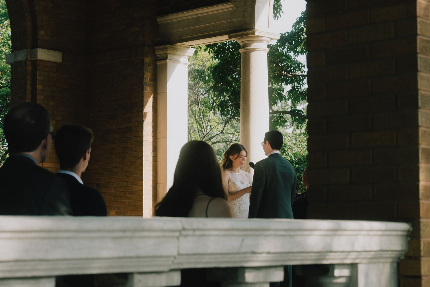 An elegant outdoor wedding ceremony takes place under white stone pillars surrounded by lush greenery.