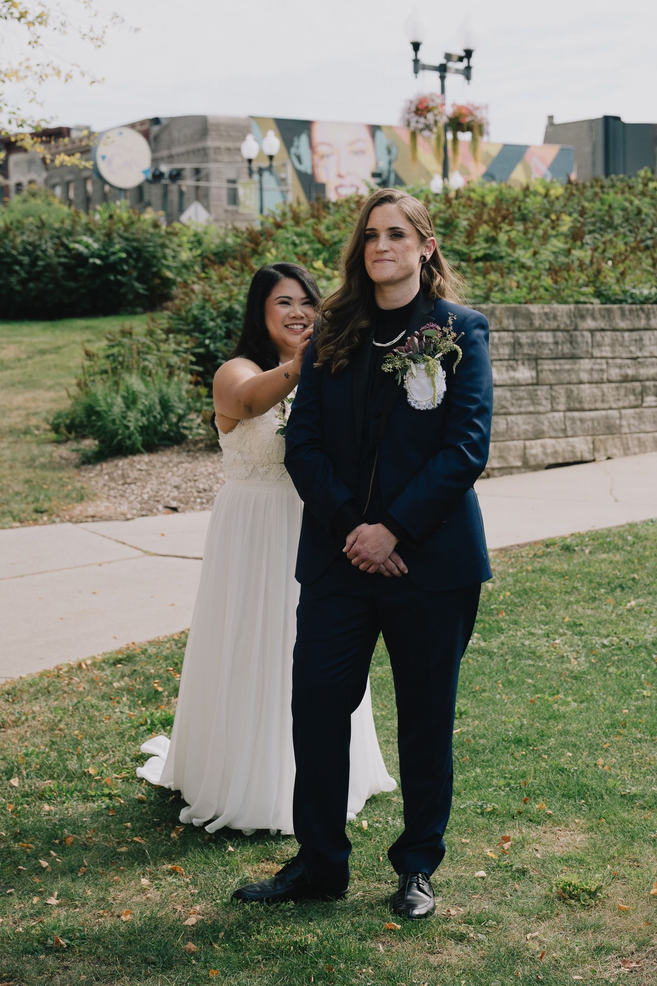 A joyful queer lesbian couple sharing a moment on their wedding day, with one in a white dress and the other in a navy suit.