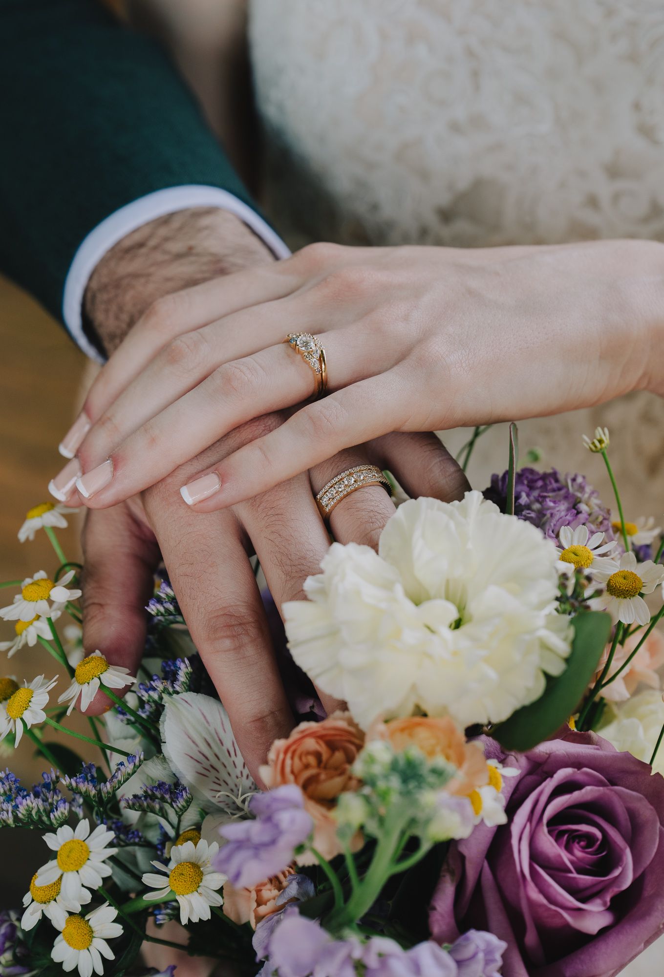 The hands of a bride and groom with wedding rings rest together on a colorful bouquet with daisies, roses, and purple flowers.
