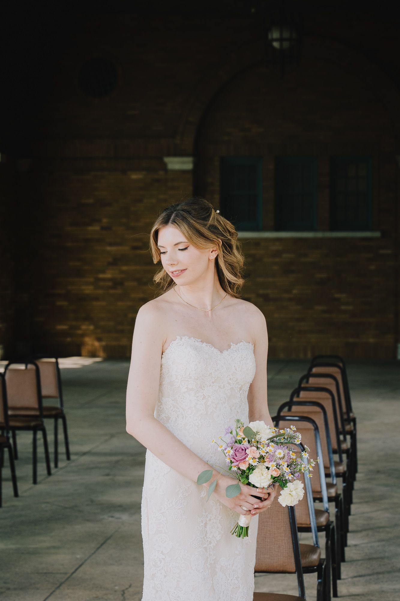 A bride in a lace wedding dress holds a delicate pink and white floral bouquet in an outdoor ceremony setting.