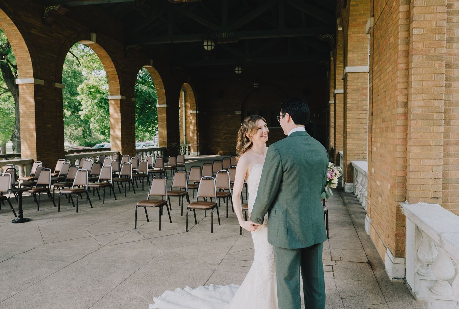 Elegant bride and groom embrace at outdoor venue with brick arches and garden backdrop.