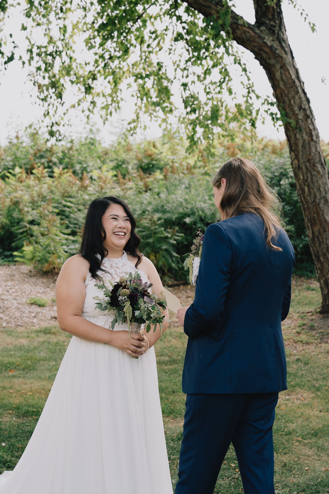 A joyful queer lesbian couple sharing a moment on their wedding day, with one in a white dress and the other in a navy suit.