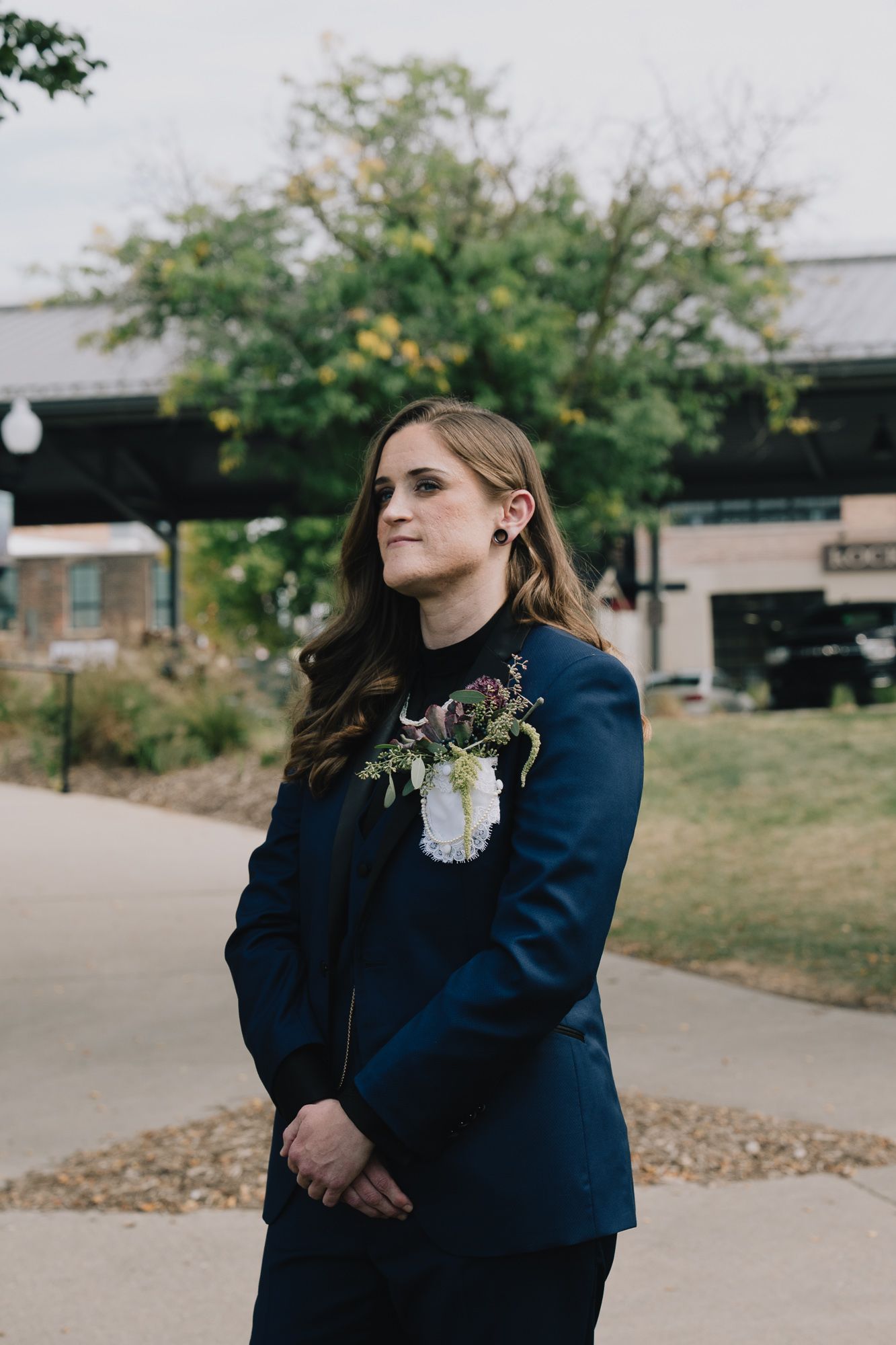 A queer lesbian woman wearing navy blue blazer stands outdoors near walkway and autumn trees on cloudy day.