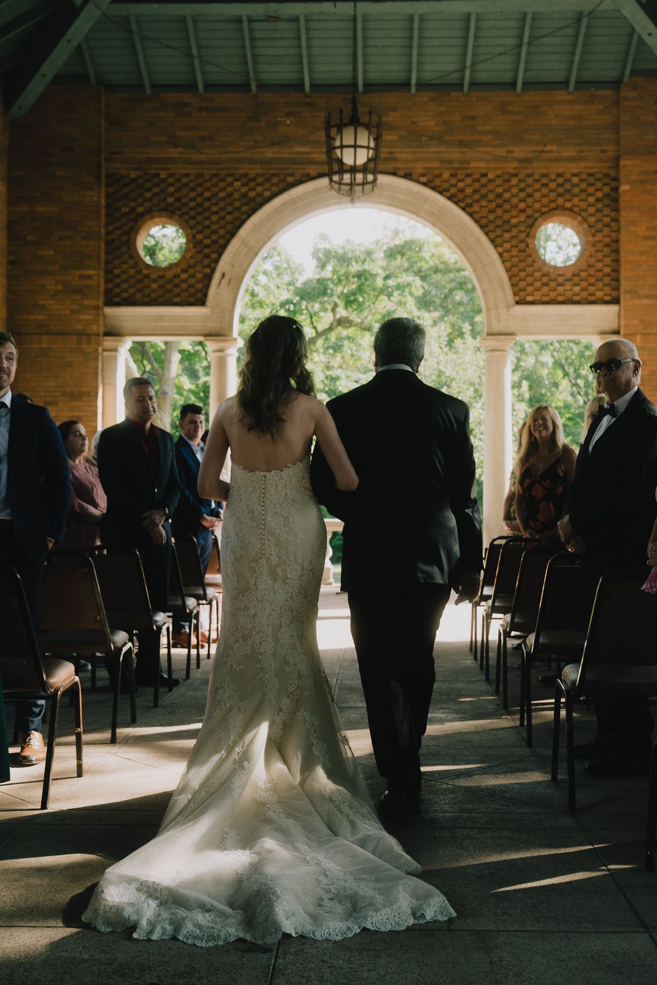 A wedding ceremony in an elegant brick archway venue with guests seated in rows as the bride walks down the aisle with her father.