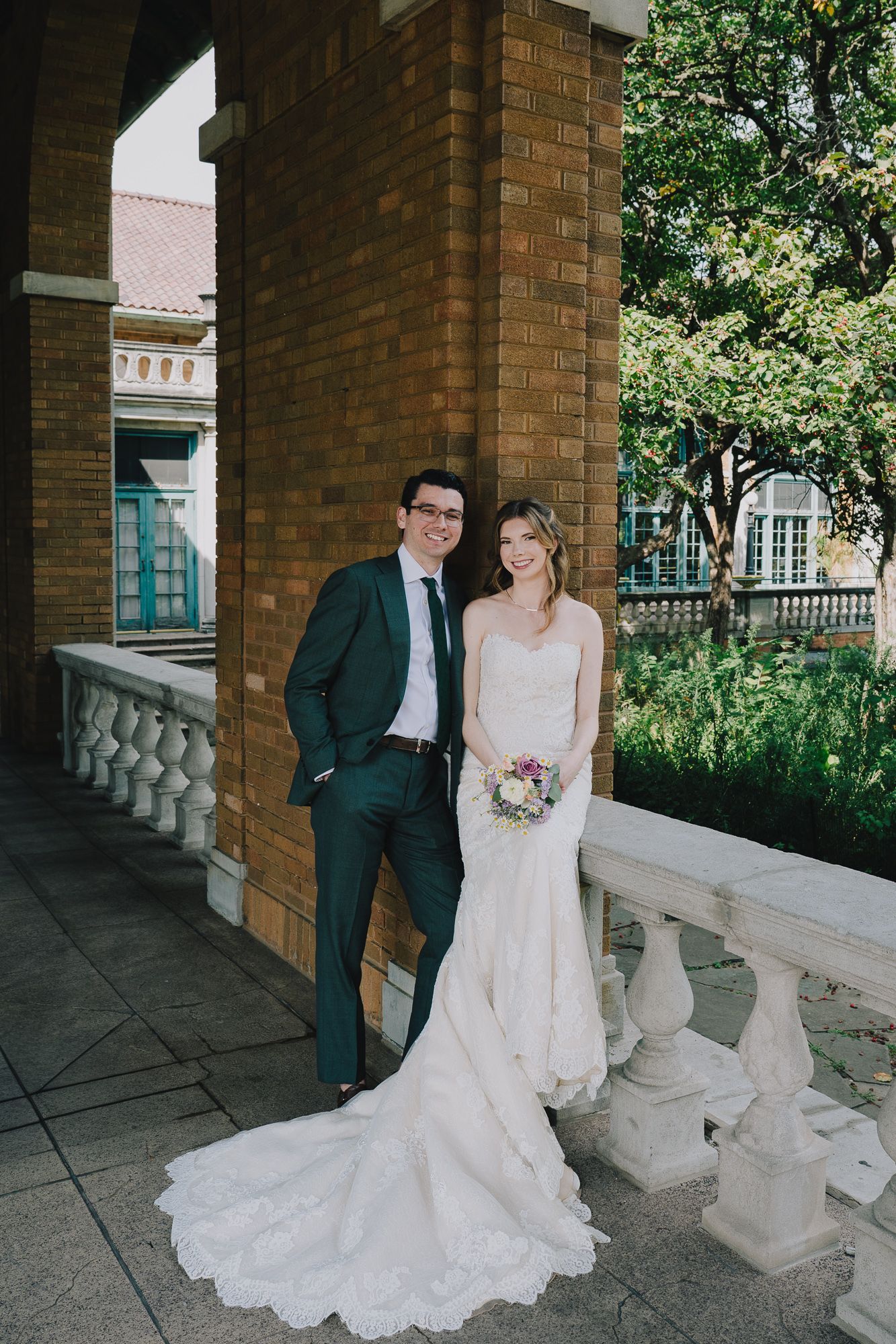 Wedding couple poses by stone balustrade in garden setting with brick columns visible in matching attire.