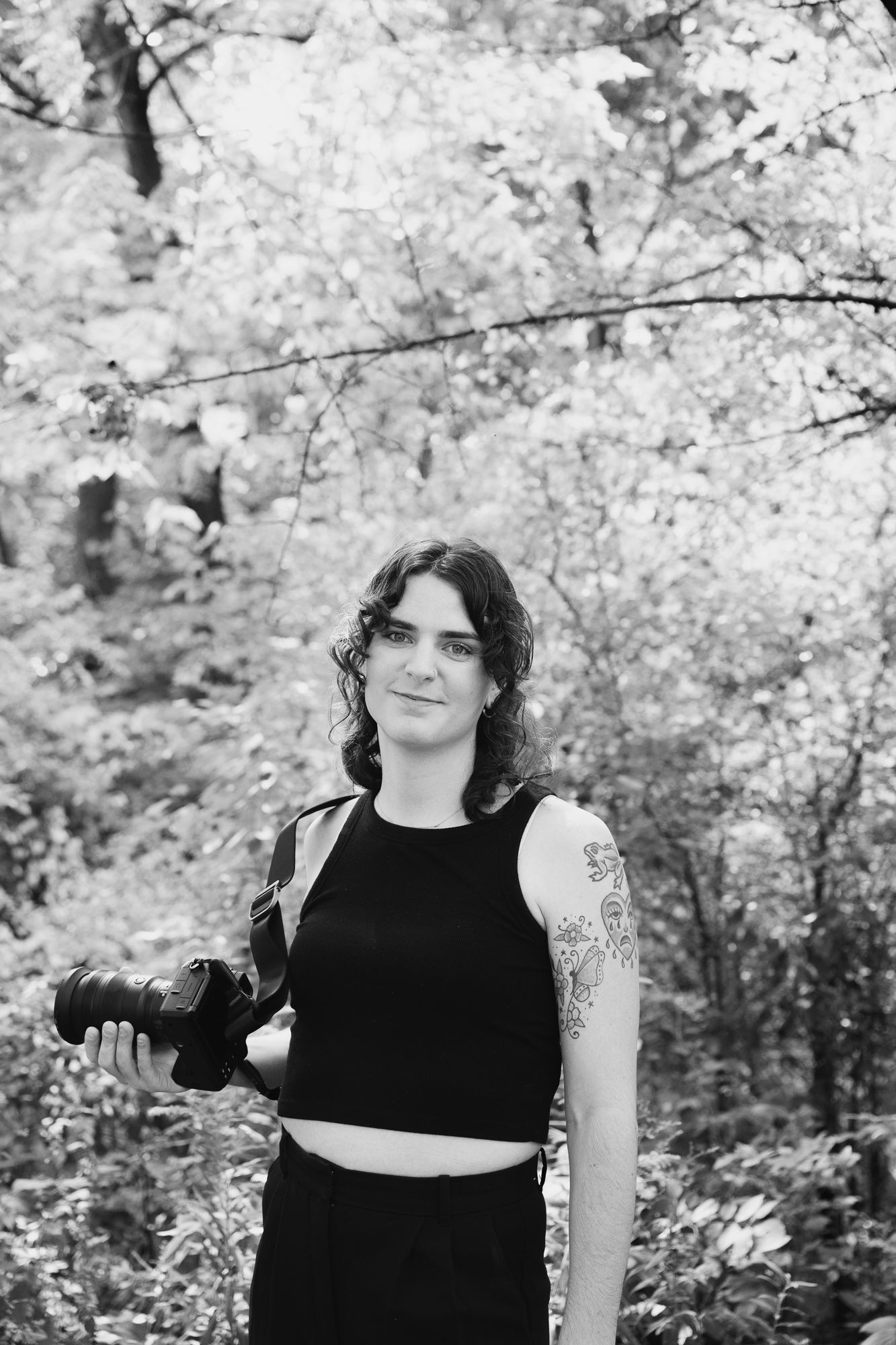 A woman wearing a black sleeveless top holding a camera poses outside among flowering trees in a black and white photograph.