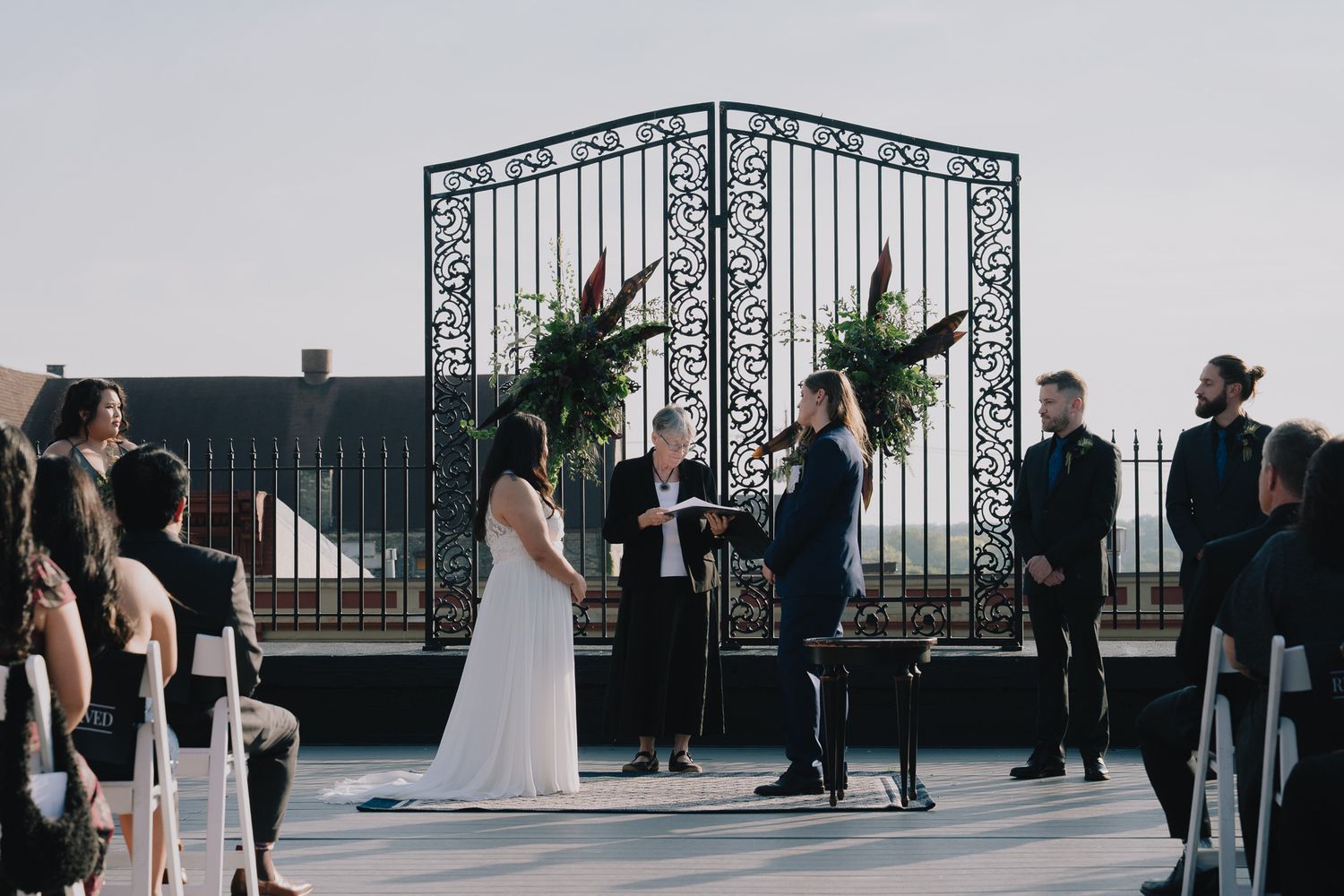 A queer lesbian couple stands on an outdoor balcony with ornate iron railings and city views in the background for their wedding ceremony.