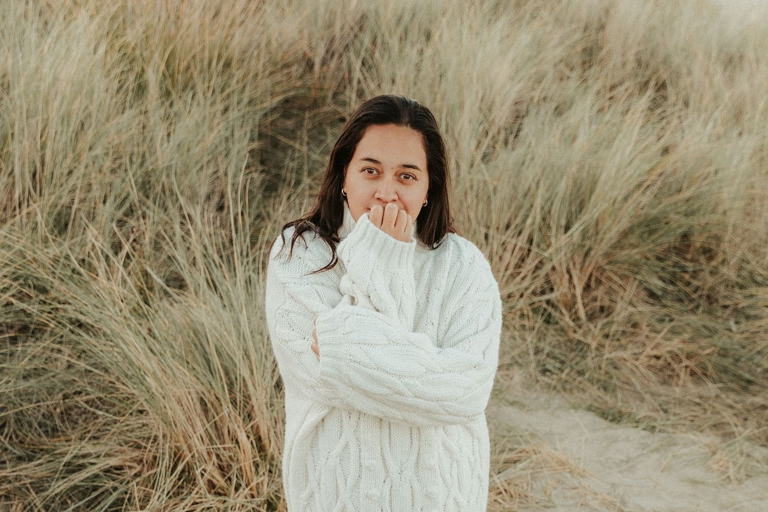 Person in cozy white sweater sitting among tall dried grass on hillside.