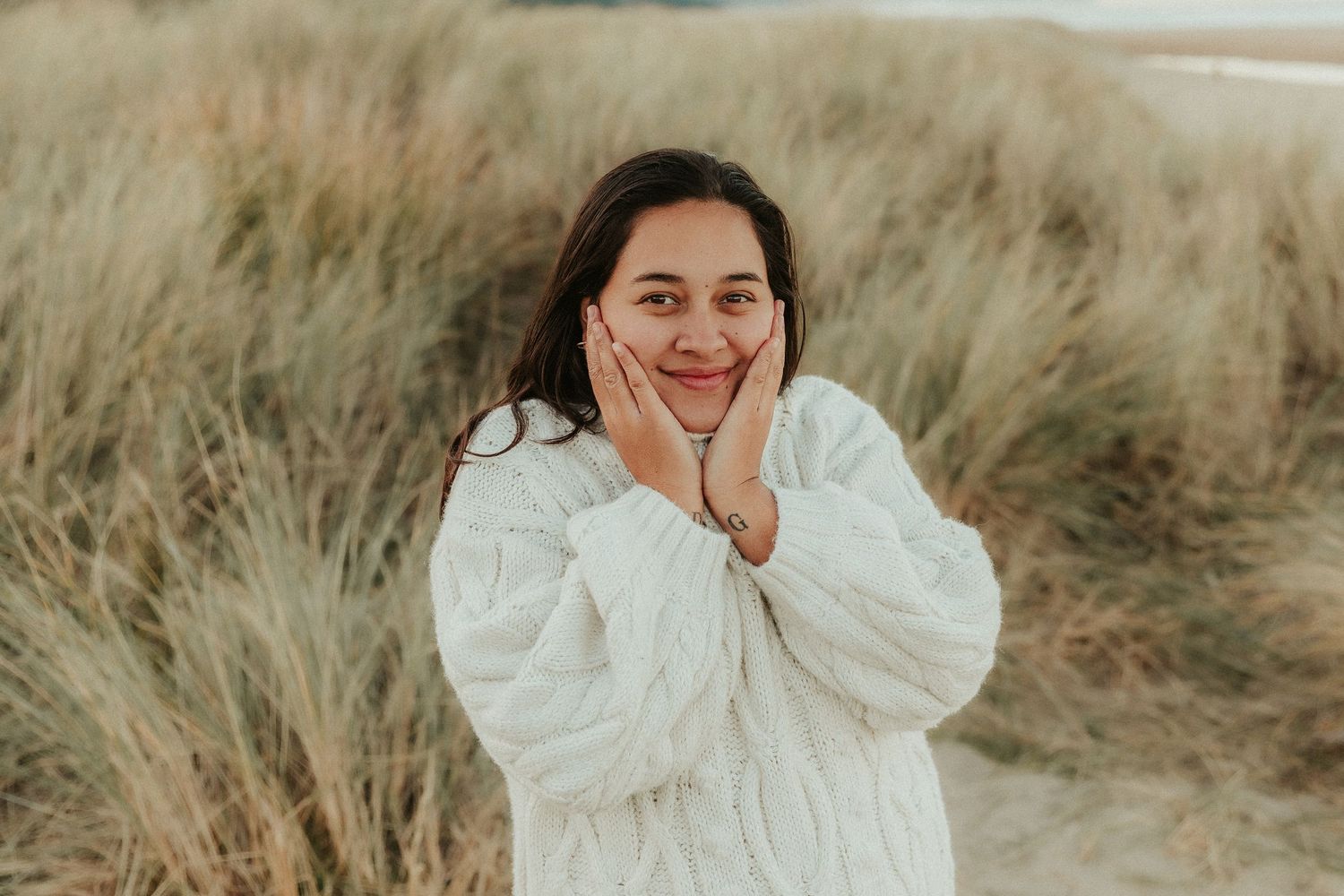 Person in white sweater poses expressively against backdrop of coastal dunes.