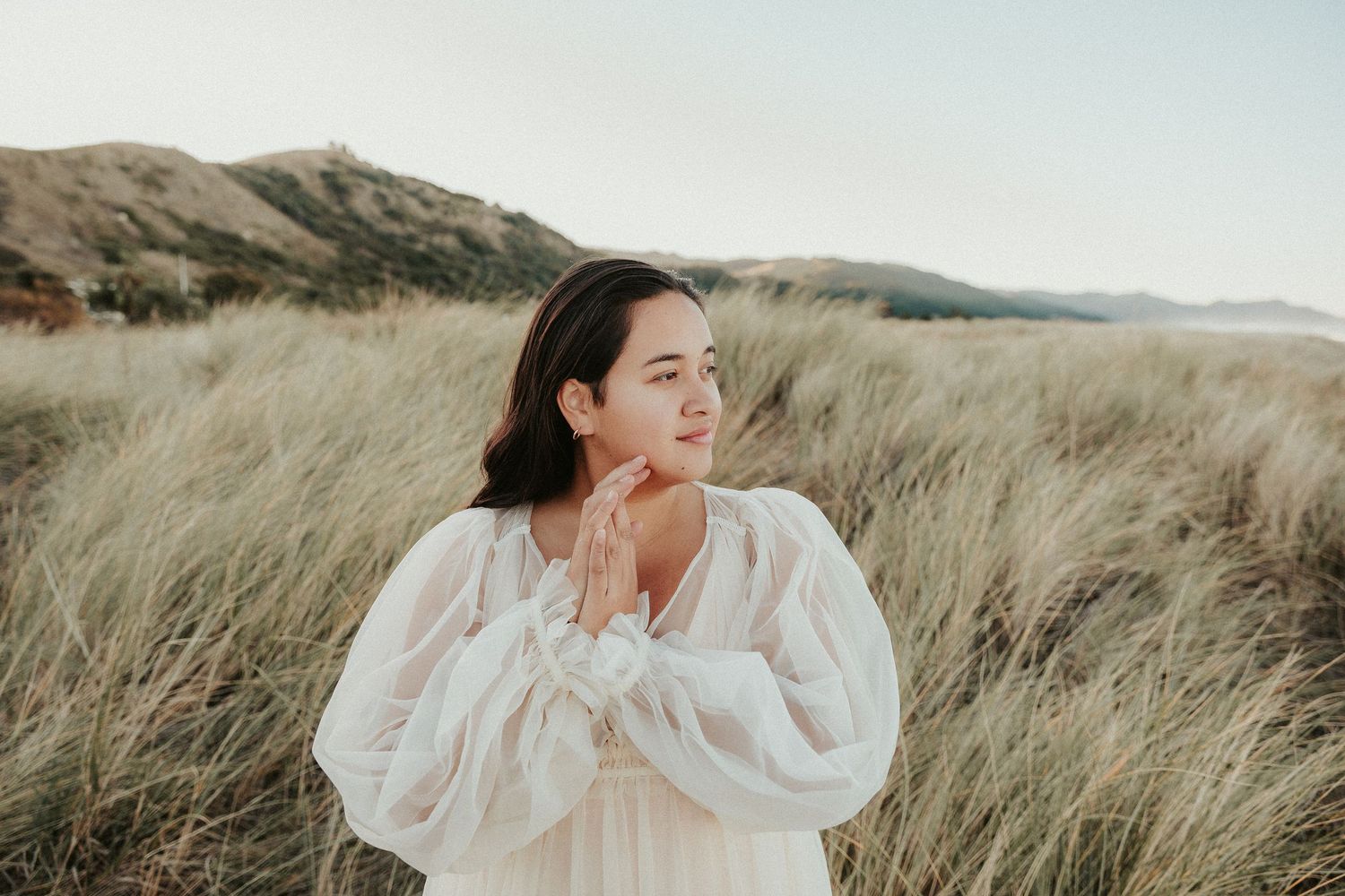 Ethereal portrait in white dress amidst windswept grass field with mountains in background.