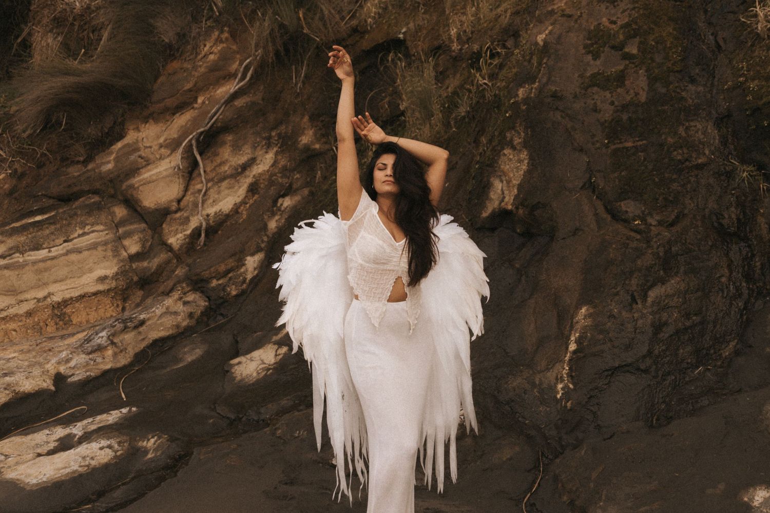 An ethereal figure in a white dress and angel wings poses gracefully against a dramatic rocky cave backdrop.
