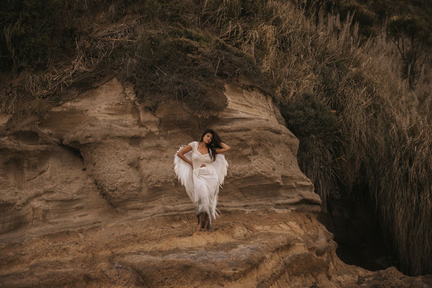 A figure in a flowing white dress stands gracefully on rugged sandstone cliffs against an earthy backdrop.