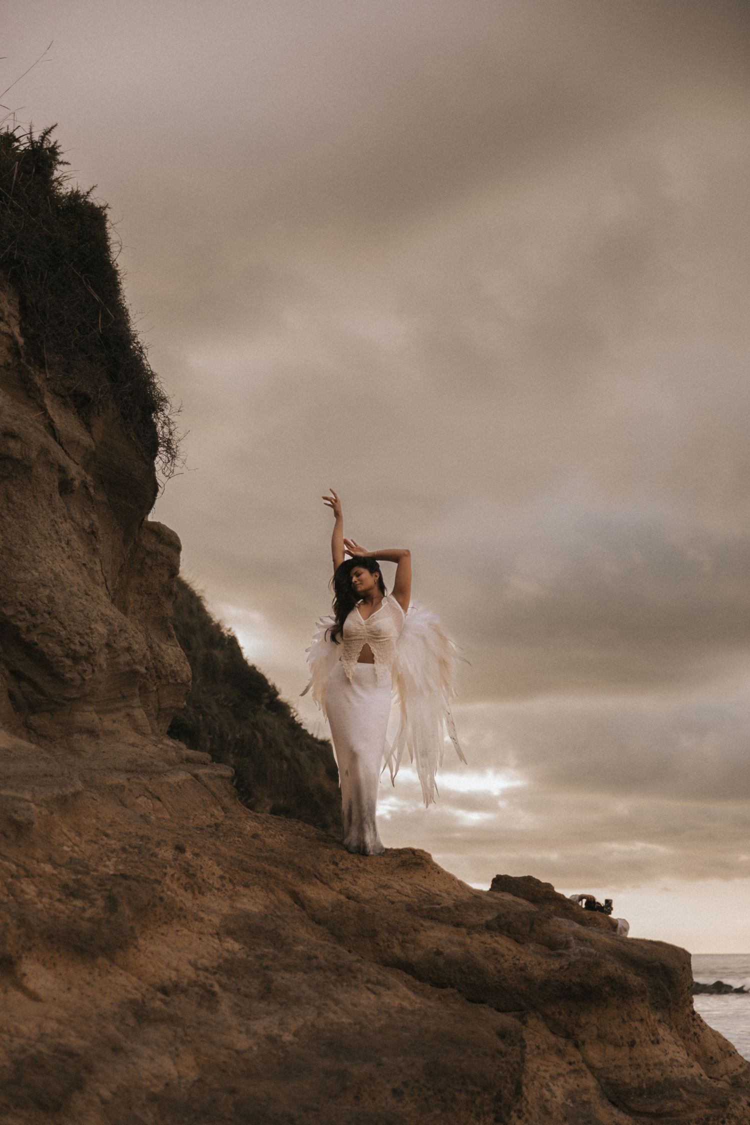 A figure in white flowing fabric poses gracefully against a dramatic coastal cliff at sunset.