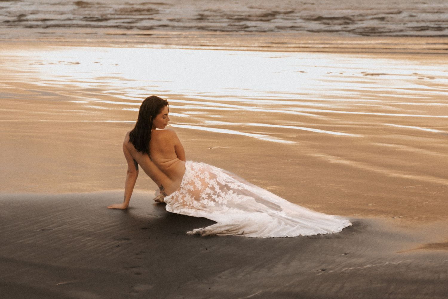 Figure in white dress lying gracefully on wet sand as waves roll in during sunset.