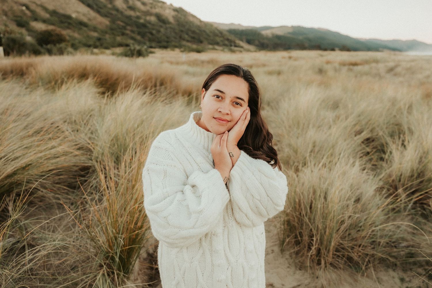 Person in white sweater poses thoughtfully in golden field at sunset.