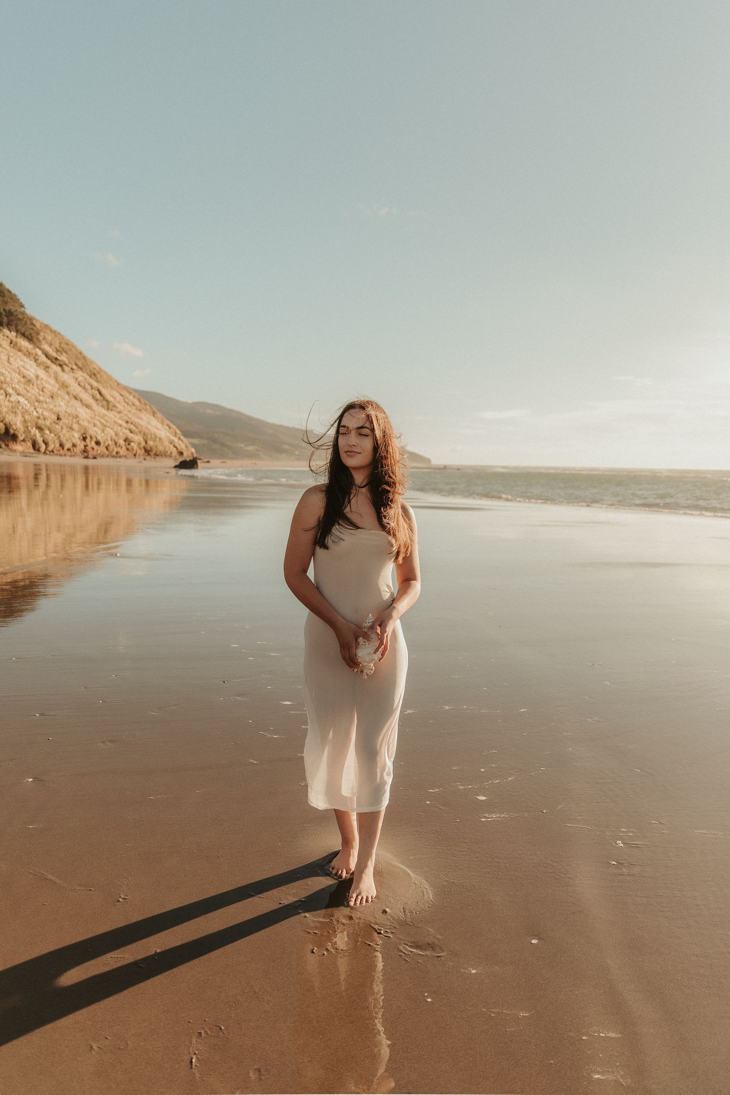 A figure in a flowing white dress walks along a serene beach at sunset with cliffs in the background.
