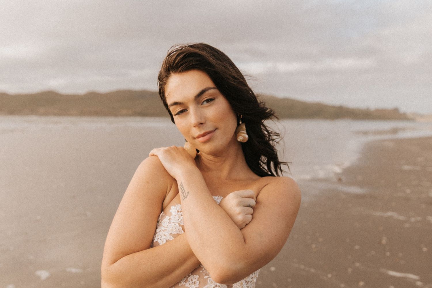 Woman in delicate lace dress stands against cloudy beach backdrop.