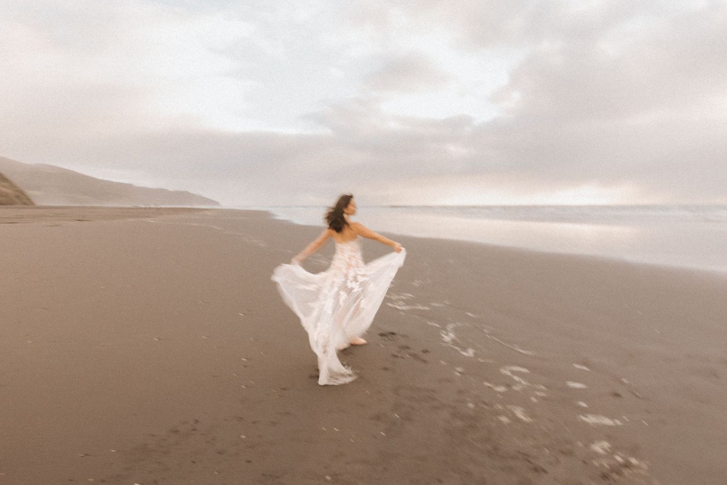 Dreamy sepia-toned photo of bride twirling on vast empty beach.