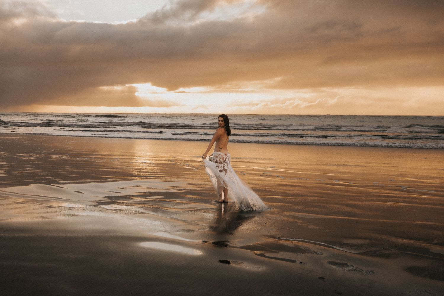 Dramatic sunset beach scene with mountains and flowing white dress on dark sand.