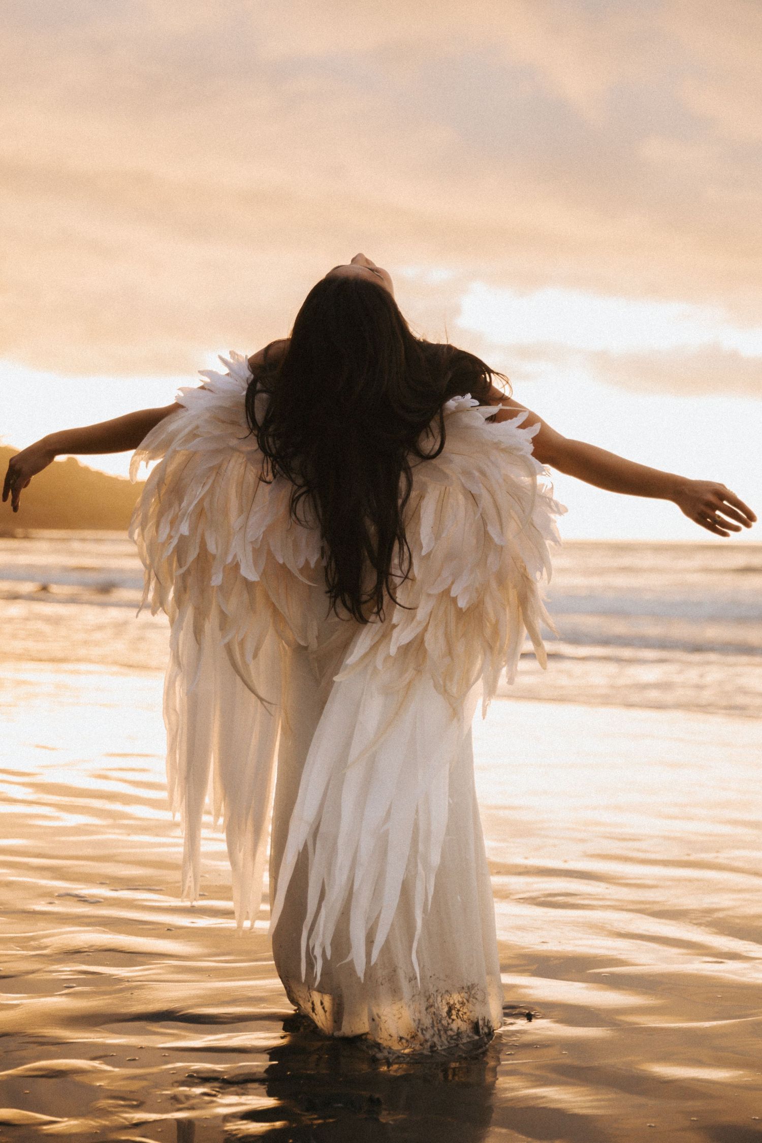 A figure in a white feathered outfit stands in shallow water at sunset creating a dreamy silhouette against golden light.