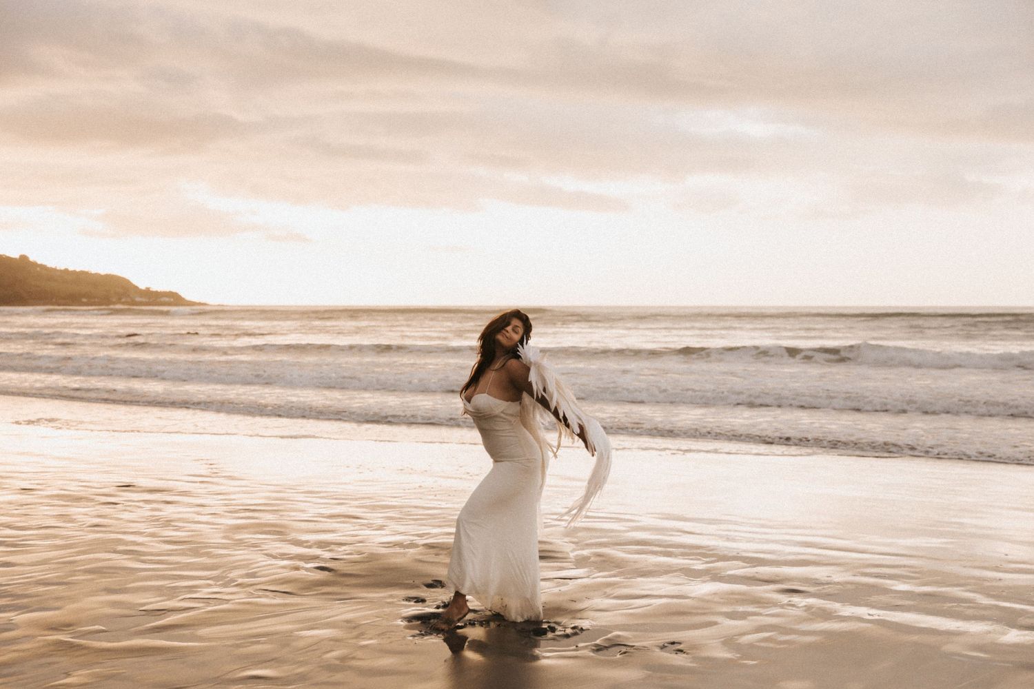 A person in a white dress walks along a misty beach shoreline at sunset creating a dreamy coastal scene.