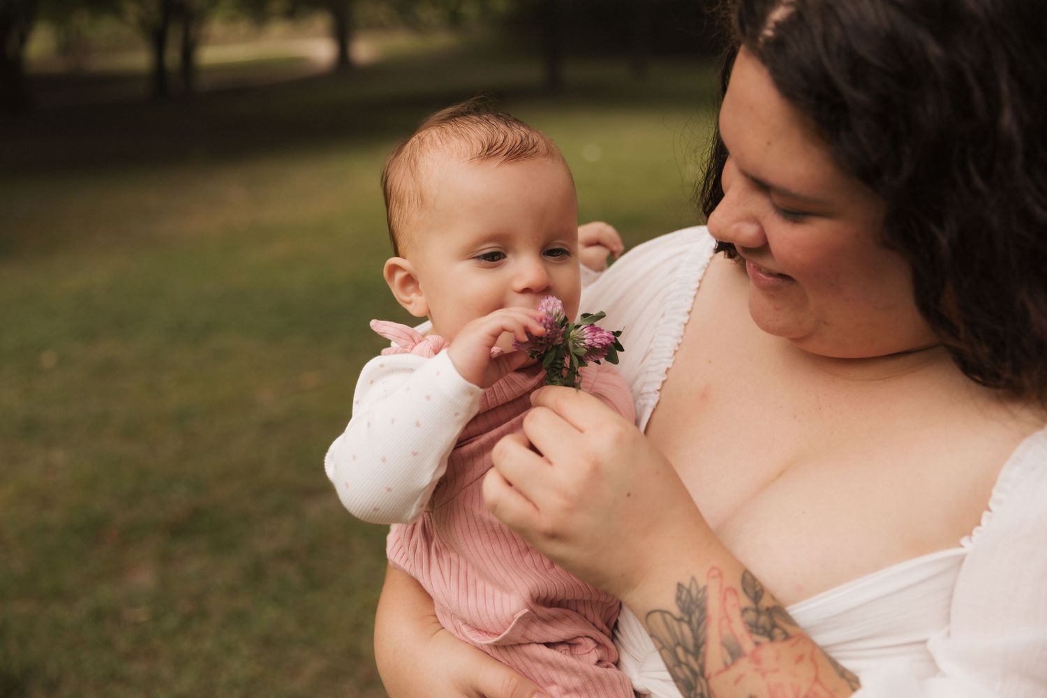 A mother holds her baby girl in a pink dress outdoors, as the infant smells a small pink flower.