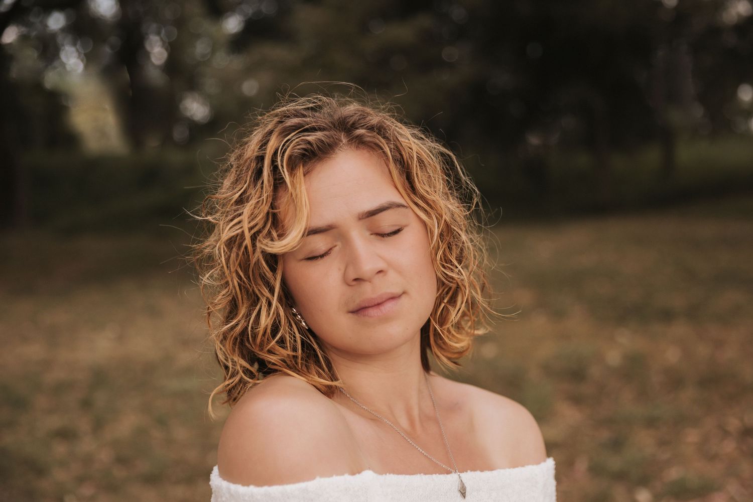 Woman in white off-shoulder top with curly hair poses with closed eyes in outdoor setting.