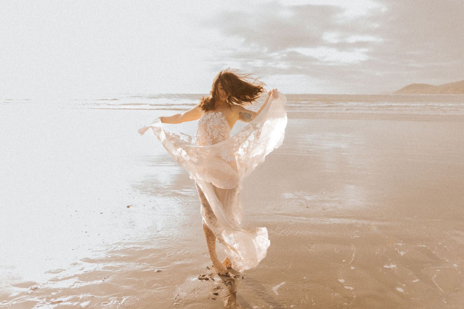 Ethereal beach portrait with flowing white dress catching wind against cloudy sky.