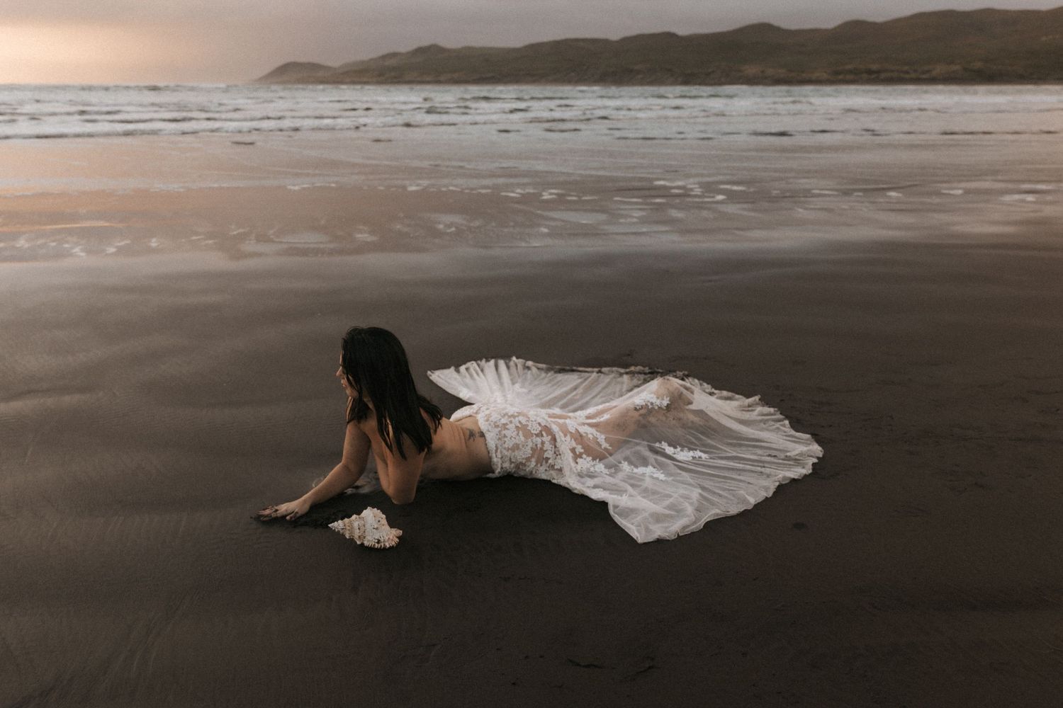 Coastal photography featuring white dress against dark sand and mountain backdrop.