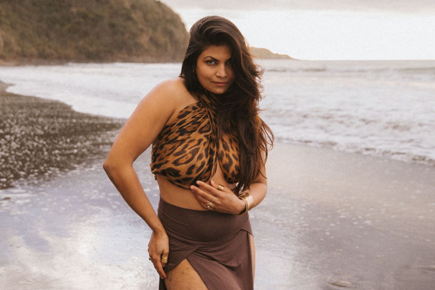 A woman in a brown dress poses gracefully on a rocky beach during sunset.