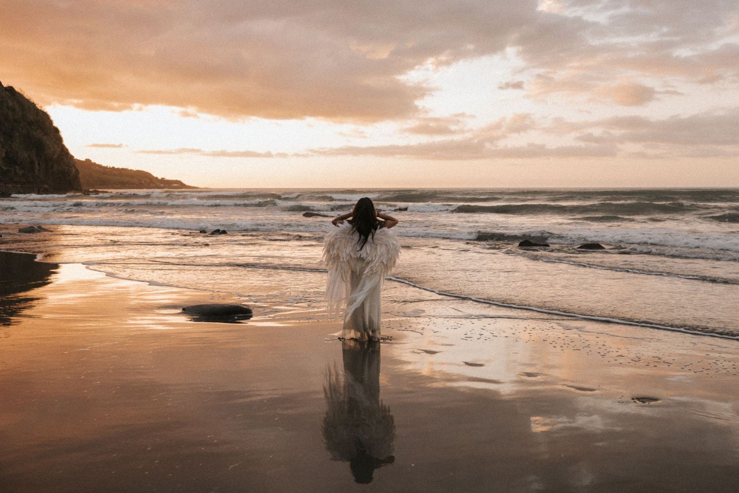 A silhouette stands on a misty beach at sunset with waves crashing on the shore and golden light reflecting on wet sand.