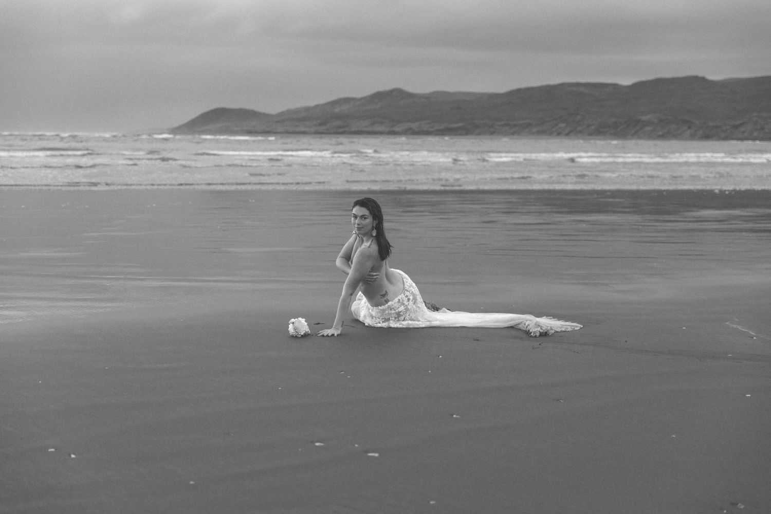 Black and white coastal scene with mountains and figure in flowing dress on beach.