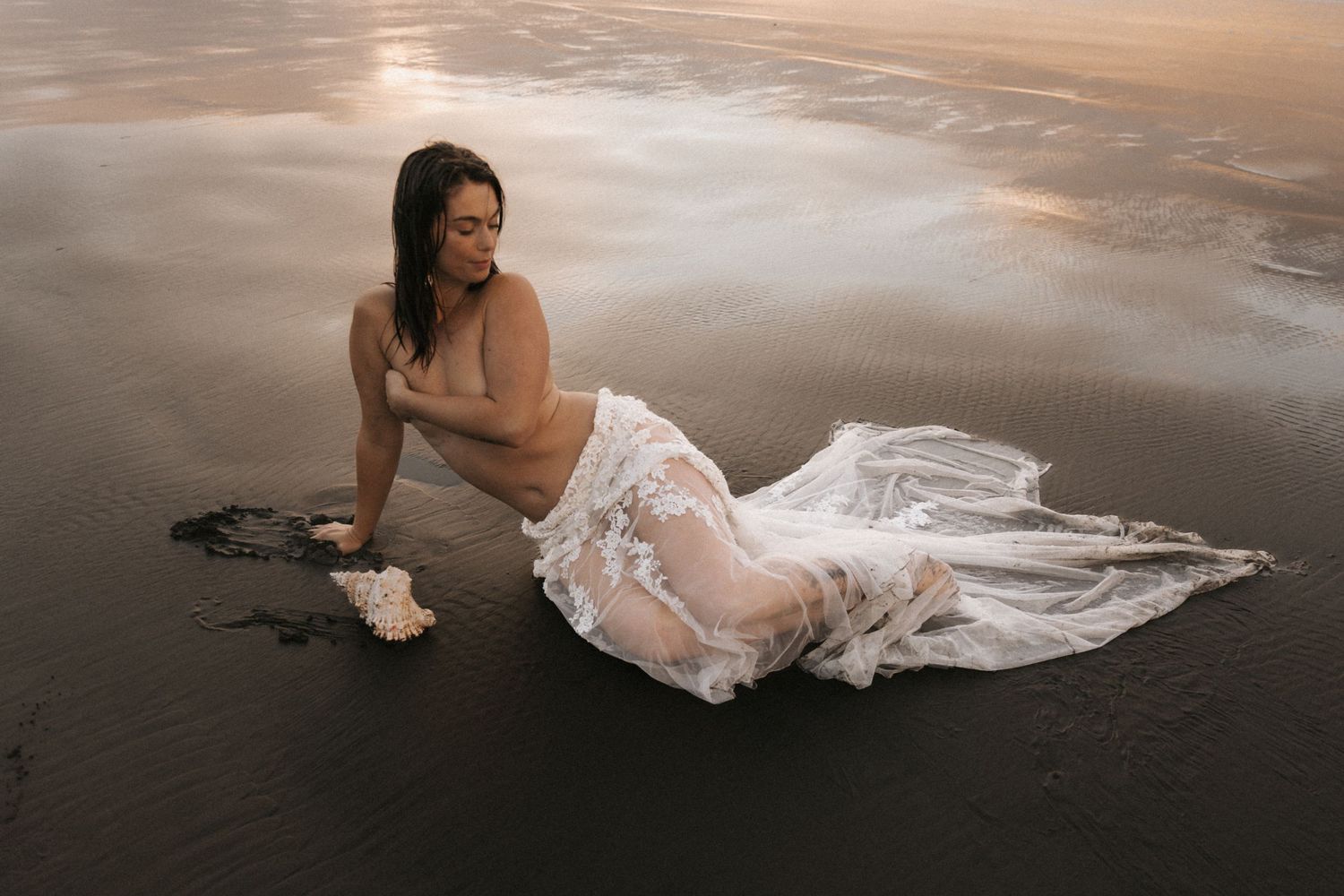 White wedding gown spreads elegantly across wet sand during golden hour at the beach.
