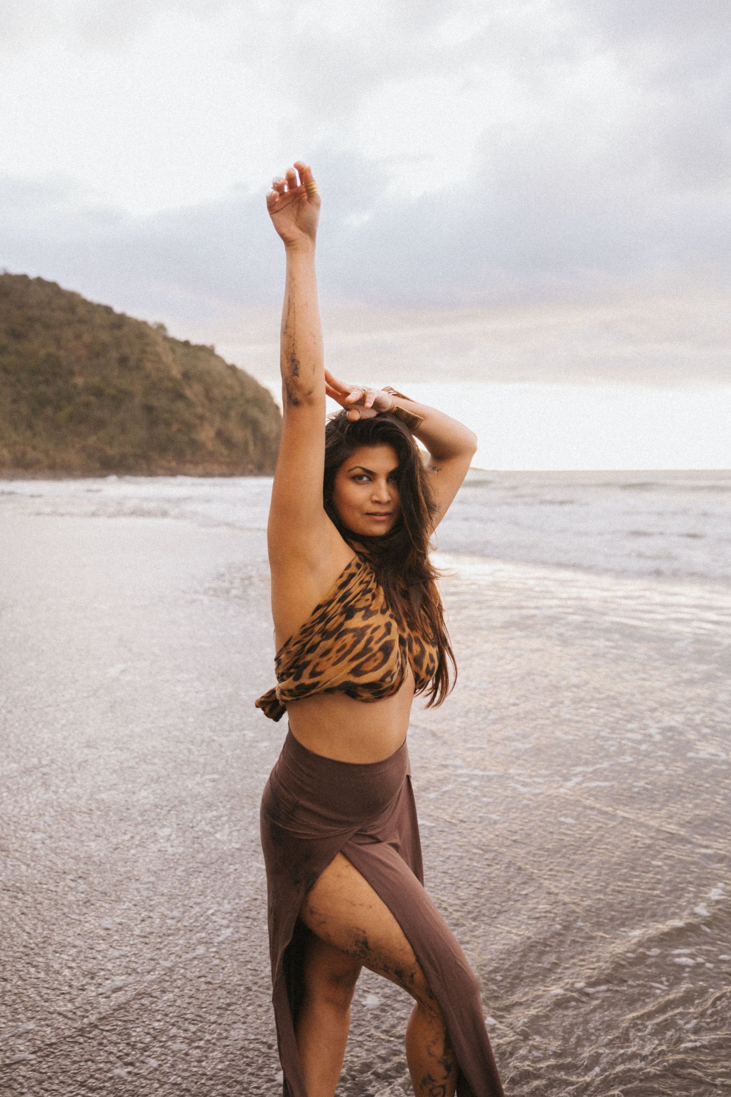 Woman in brown athletic wear poses gracefully on beach during sunset while waves crash on shore.