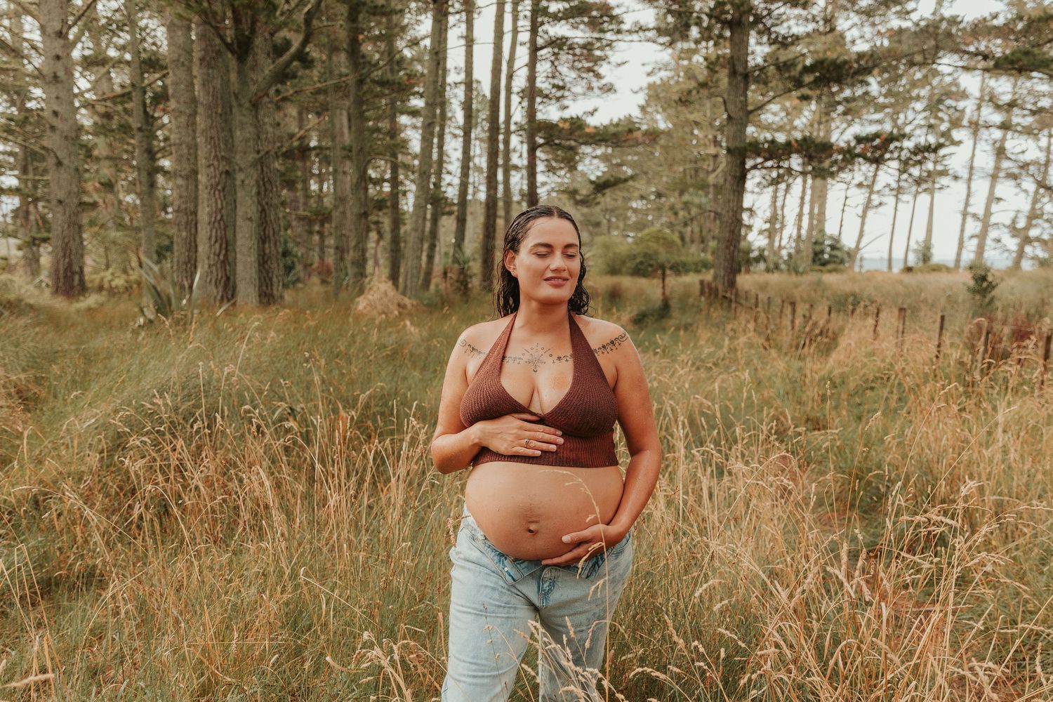 A series of maternity photos showing a pregnant woman in a brown top and jeans posing thoughtfully ion a rainy day in the forest.