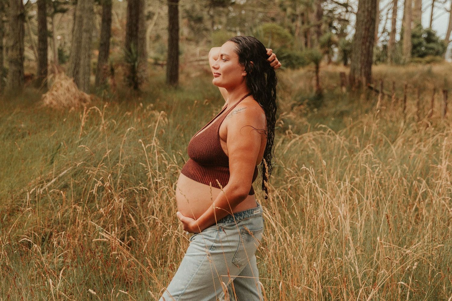 Pregnant woman in jeans walks through a golden autumn field at sunset with tall trees in the background.