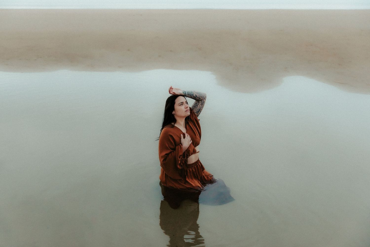 Figure in a brown coat poses artistically while standing in shallow reflective water.